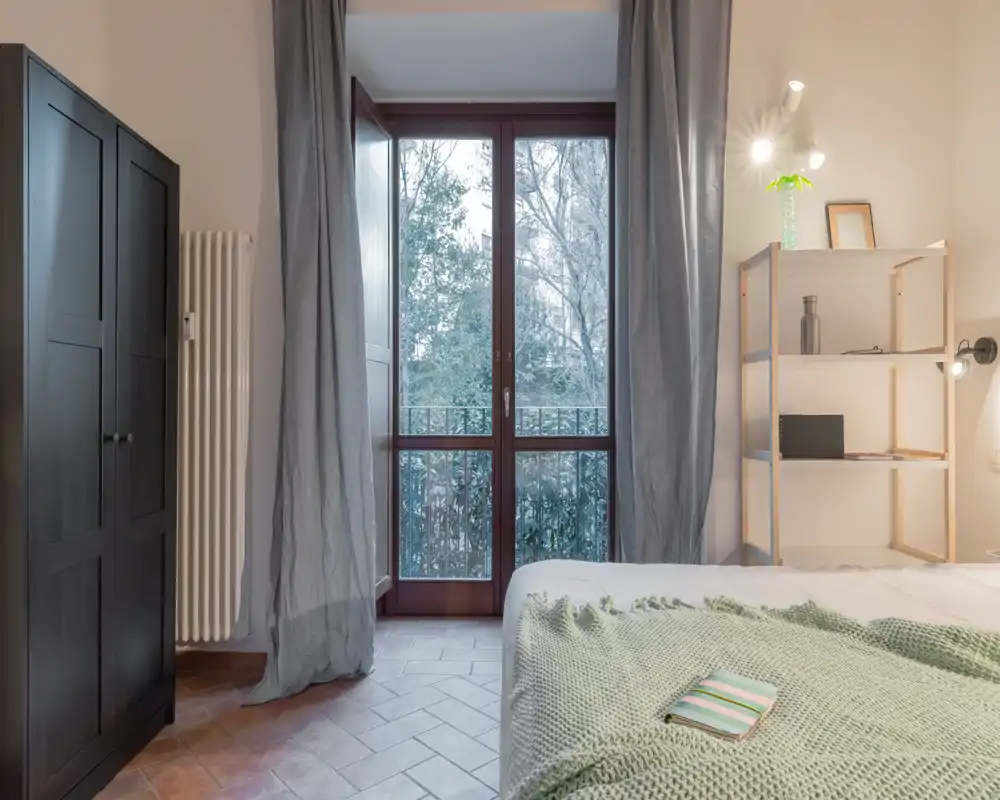 Camera da letto luminosa e ben arredata con ampia finestra/porta sul balcone, tessili morbidi e una scaffalatura minimale — perfetta per un rifugio tranquillo con molta luce naturale.