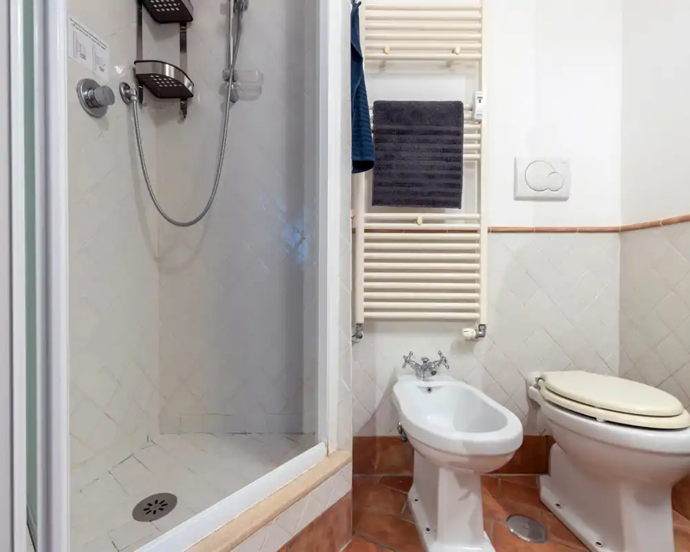 Bagno compatto e ben illuminato con doccia piastrellata, bidet e WC; pavimento in cotto e piastrelle bianche a parete per un aspetto pulito e tradizionale.