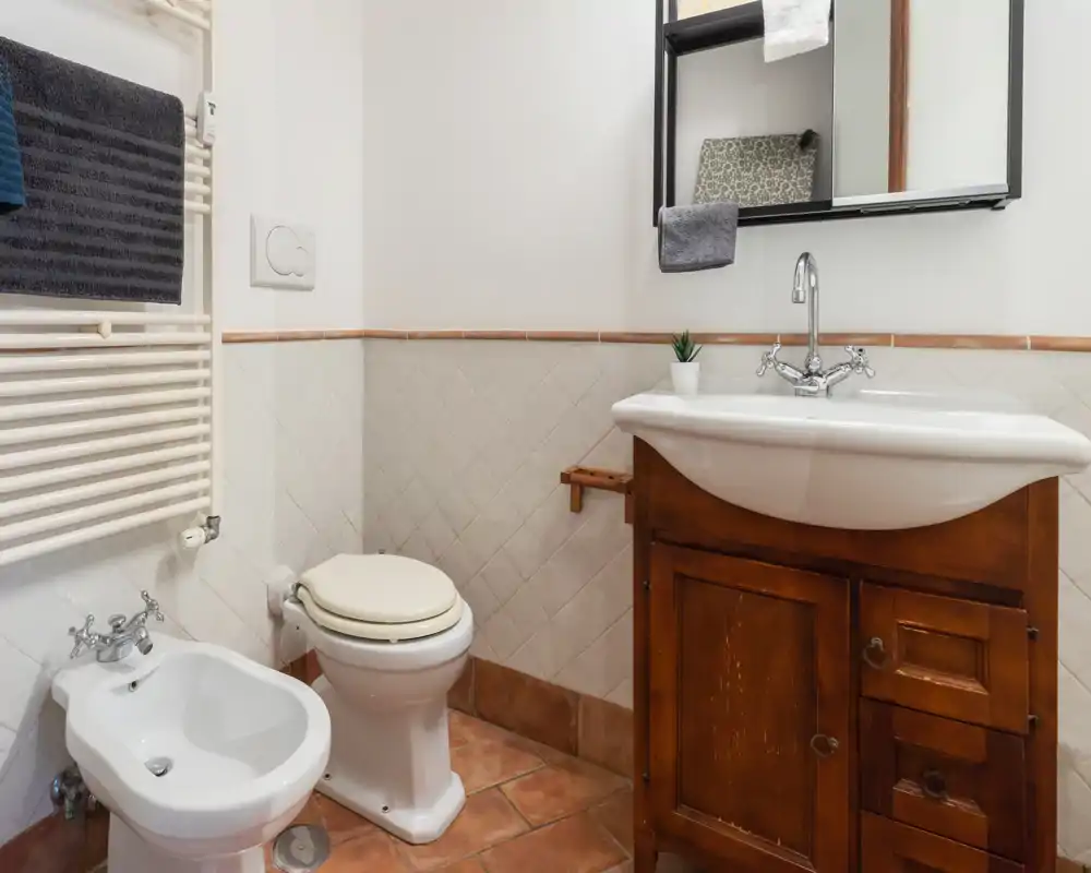 Bagno compatto e ben curato con WC, bidet e mobile lavabo in legno; piastrelle chiare e specchio rendono l'ambiente pulito e luminoso.