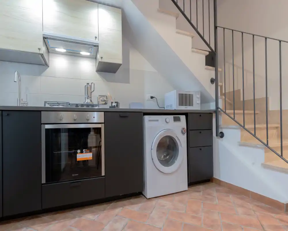 Cucina moderna compatta sotto la scala con forno incassato, lavello, lavatrice e microonde; pavimento in cotto che aggiunge calore.