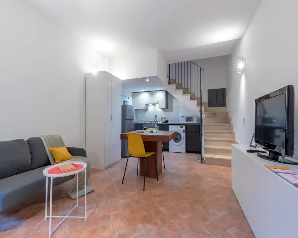 Zona giorno open-space con angolo cottura sotto le scale. L’immagine mostra divano, tavolo da pranzo con sedie, mobile TV e elettrodomestici visibili (frigorifero, lavatrice) — ideale per un appartamento urbano accogliente.