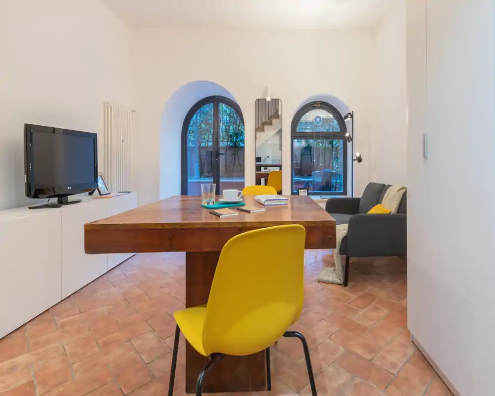Area pranzo calda e luminosa con un grande tavolo in legno e sedie gialle moderne; ampie finestre ad arco portano luce naturale e sullo sfondo si vede un angolo soggiorno accogliente con divano.