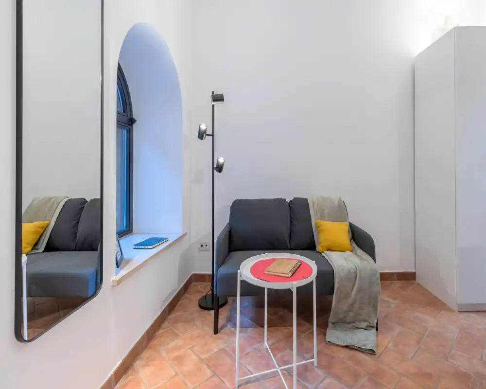 Zona giorno compatta con divano moderno per due persone, plaid e cuscino, un piccolo tavolino rotondo e una lampada da terra. Pareti bianche e pavimento in cotto donano un angolo luminoso e accogliente.
