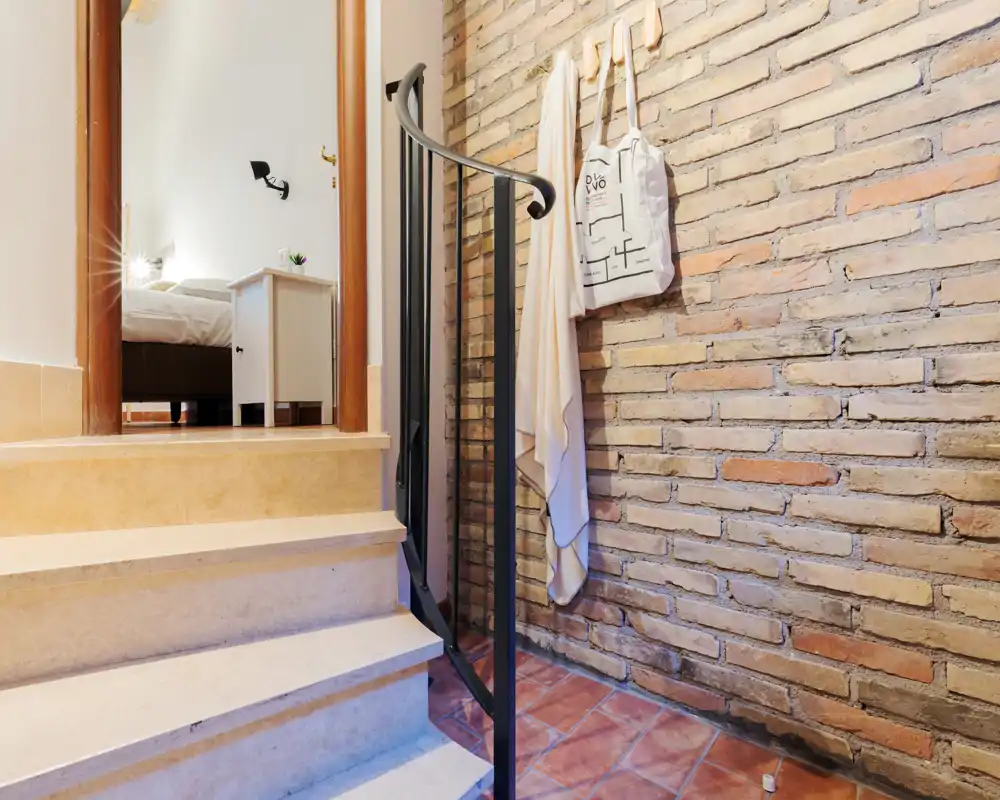 Piccolo ingresso/scale con parete in mattoni a vista calda, pavimento in piastrelle e una breve rampa che conduce a una camera da letto. Ganci decorativi e una borsa appesa aggiungono carattere; lo spazio è curato e accogliente.