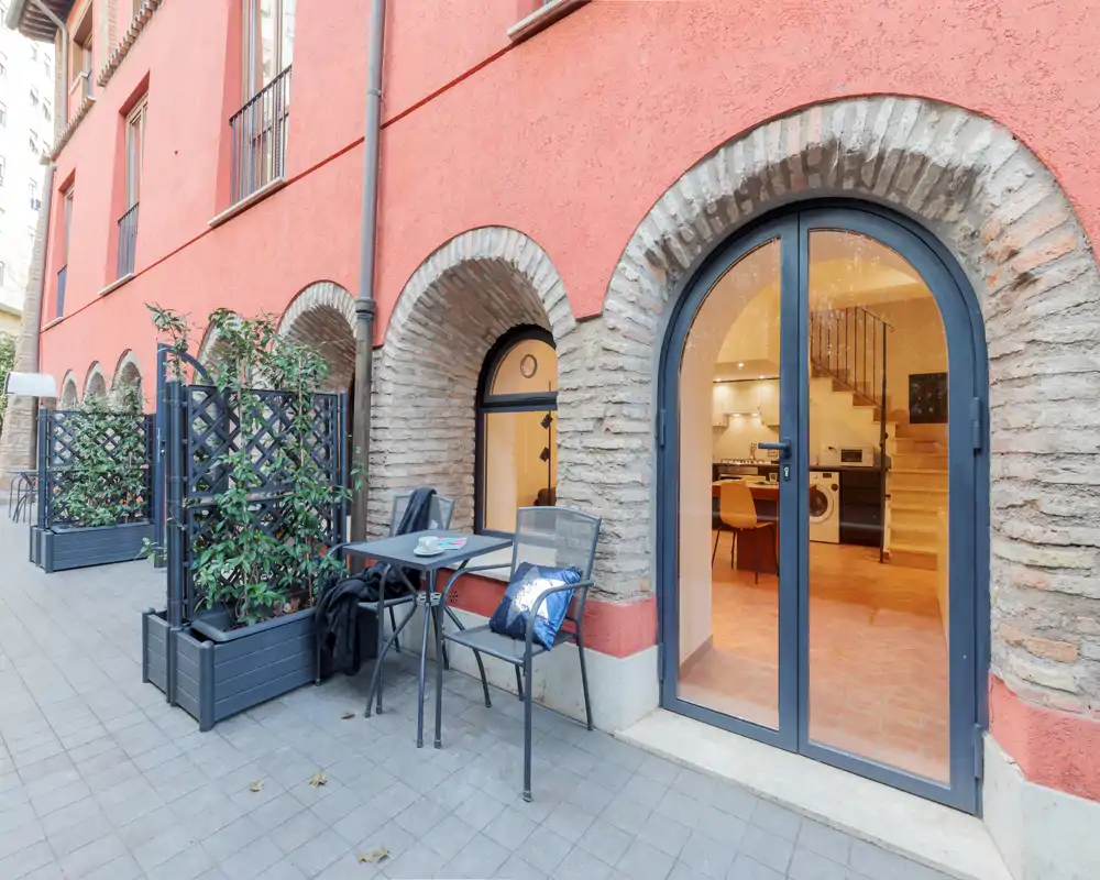 Incantevole terrazza a livello strada con un piccolo tavolo e due sedie, fioriere per la privacy e accesso diretto all'interno riscaldato da luci calde attraverso porte ad arco in vetro.