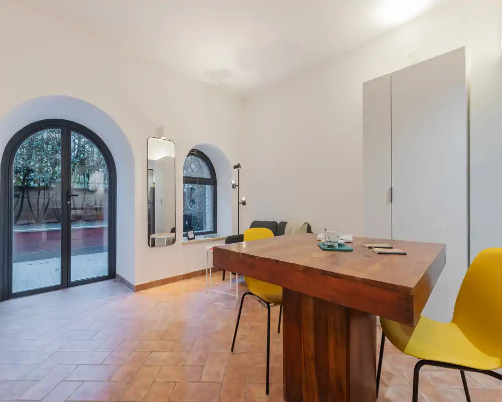 Zona pranzo luminosa e minimale con un tavolo in legno massiccio e sedie gialle moderne su pavimento in cotto, con porte ad arco che si affacciano all'esterno.