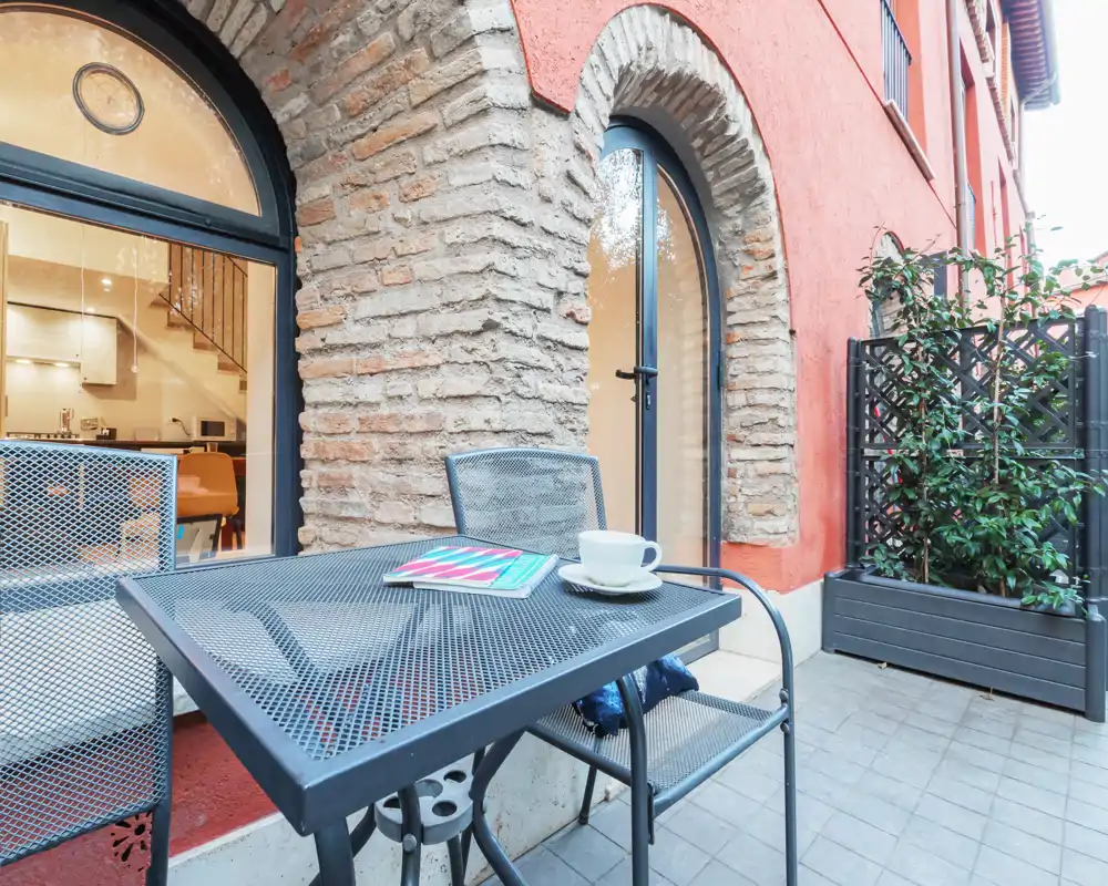 Piccola terrazza accogliente con tavolo e sedie in metallo accanto a un suggestivo arco in mattoni e piante in vaso — perfetta per un caffè mattutino o una lettura leggera.