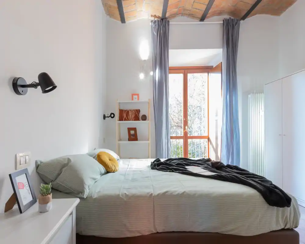 Chambre moderne et lumineuse avec un grand lit double, textiles doux et une fenêtre laissant entrer beaucoup de lumière naturelle. Le plafond en brique apparente et la décoration épurée créent une ambiance chaleureuse et stylée.