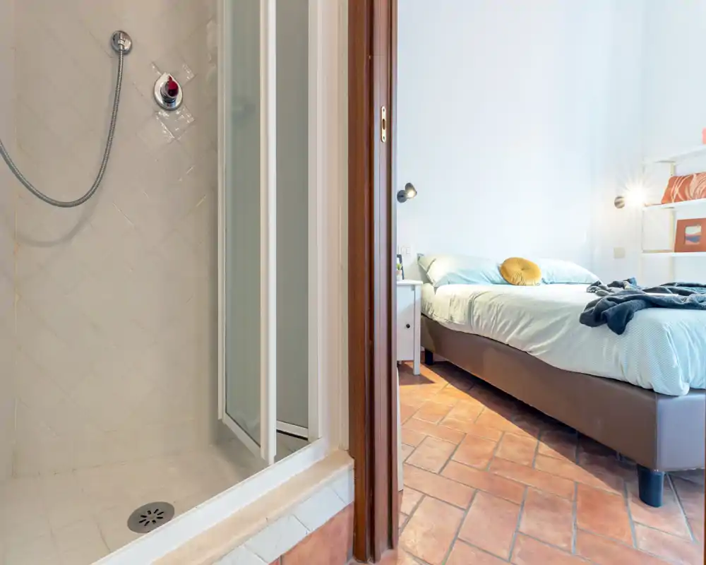 Chambre lumineuse et soignée vue à travers une porte ouverte, avec un lit double, table de chevet et sol en terre cuite chaleureux.