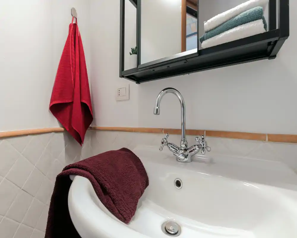 Lavabo de salle de bains lumineux et détaillé avec robinet chromé, serviette sur le lavabo et serviette rouge accrochée; demi-mur carrelé et étagère miroir au-dessus.