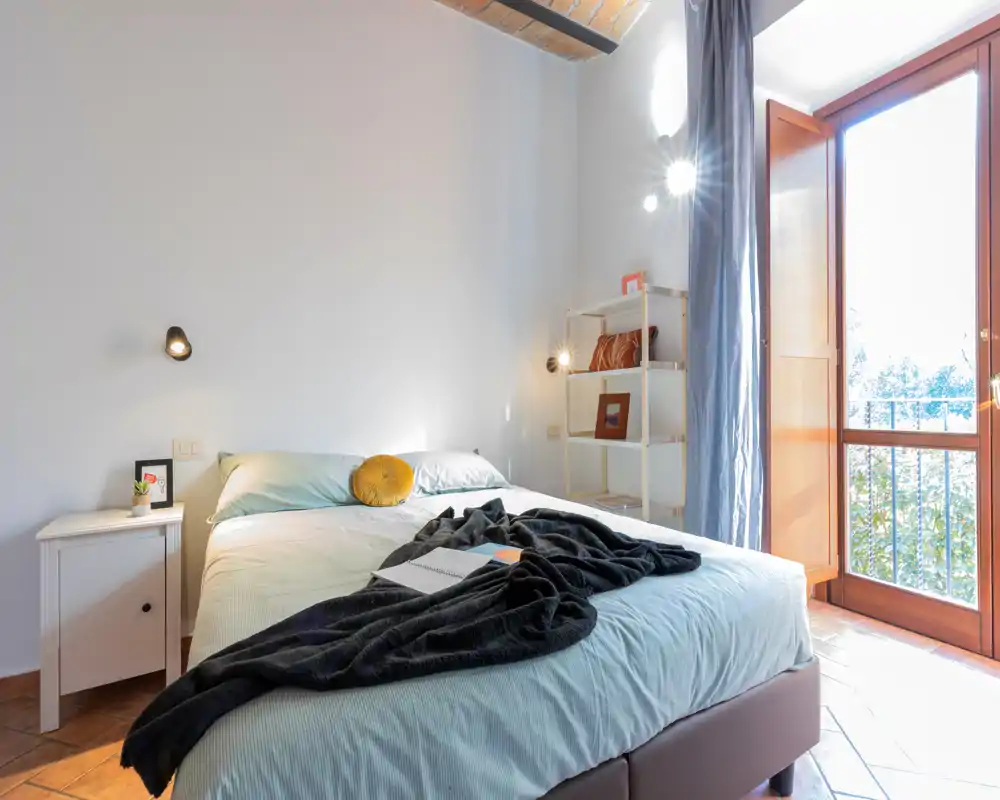 Chambre lumineuse et chaleureuse avec un lit double, linge de lit doux, coussin décoratif et plaid noir; grande porte-fenêtre donnant sur un balcon et apportant beaucoup de lumière naturelle.