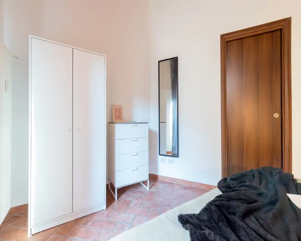 Coin de chambre minimaliste et lumineux avec le bord du lit et une couverture foncée, une armoire blanche, une commode et une grande glace — idéal pour une annonce de location moderne.