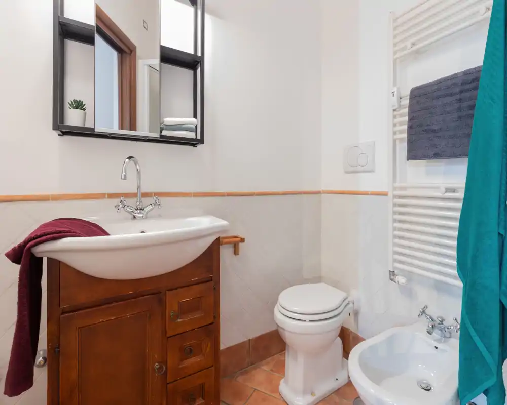 Salle de bain lumineuse avec un lavabo blanc sur meuble en bois, miroir mural, WC et bidet ; sol carrelé chaleureux et serviettes bien rangées.