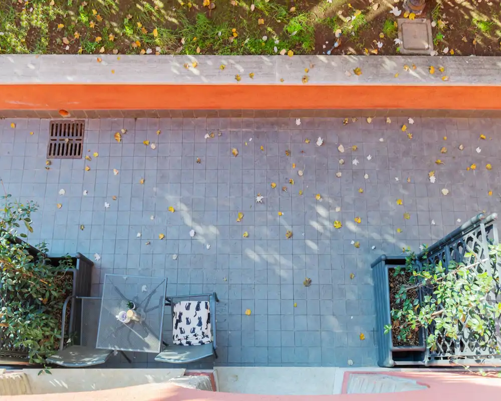 Petit balcon carrelé vu de dessus avec une table en verre, une chaise et des plantes en pot ; des feuilles d'automne éparses et une lumière douce créent un coin extérieur agréable.