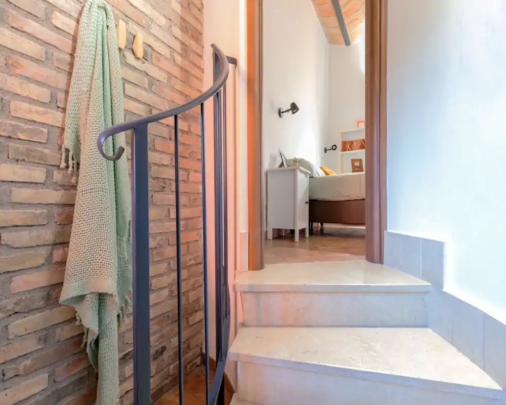 Vue sur une chambre depuis un petit escalier : une table de chevet et le lit partiellement visible à travers l'encadrement de la porte, avec des briques et des textiles doux au premier plan.
