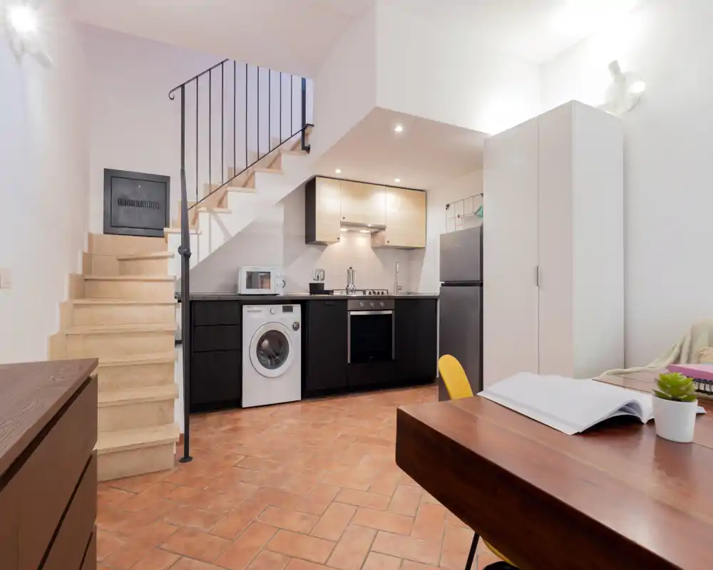 Kitchenette ouverte avec électroménagers modernes, lave-linge sous le plan de travail, réfrigérateur et petite table repas/travail; sol en terre cuite et murs neutres qui apportent chaleur et fonctionnalité.