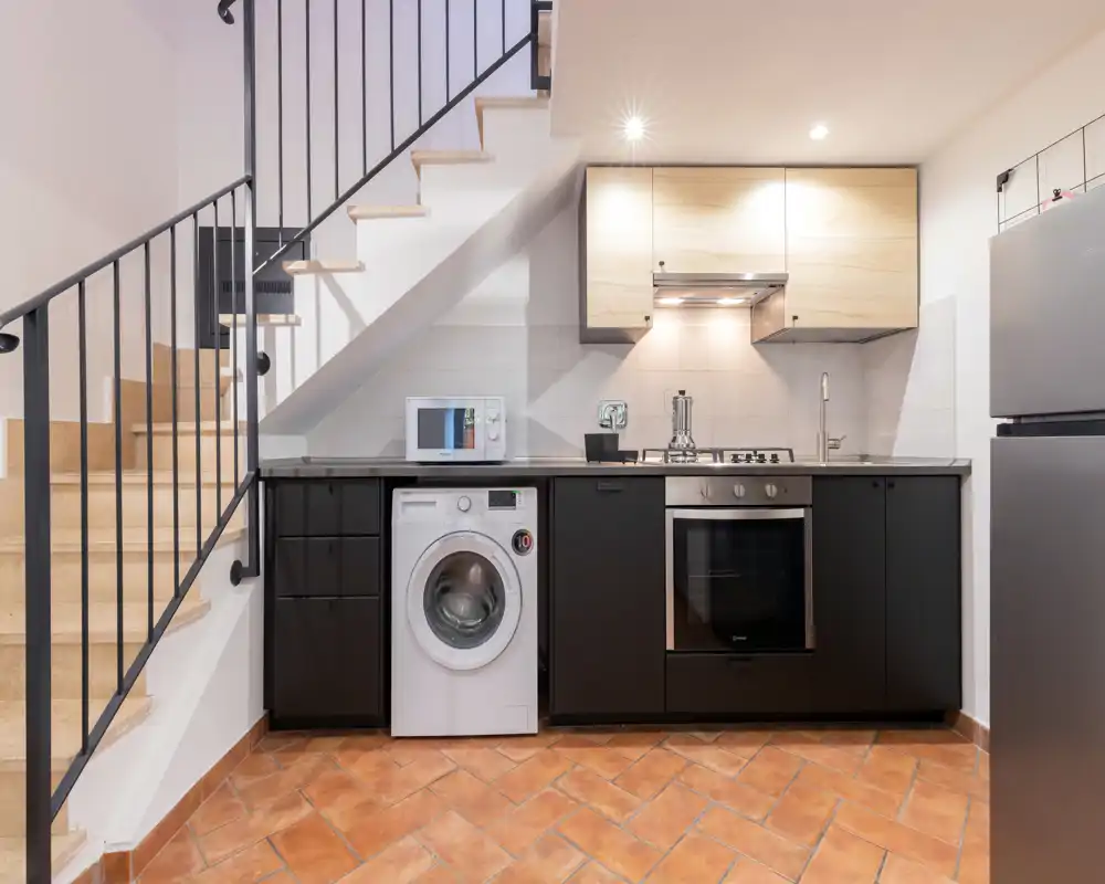 Cuisine moderne compacte sous l'escalier avec four encastré, plaque à gaz, micro-ondes et lave-linge. Meubles hauts en bois clair et éléments bas noirs contrastent avec le carrelage terre cuite, offrant un coin cuisine pratique et esthétique.