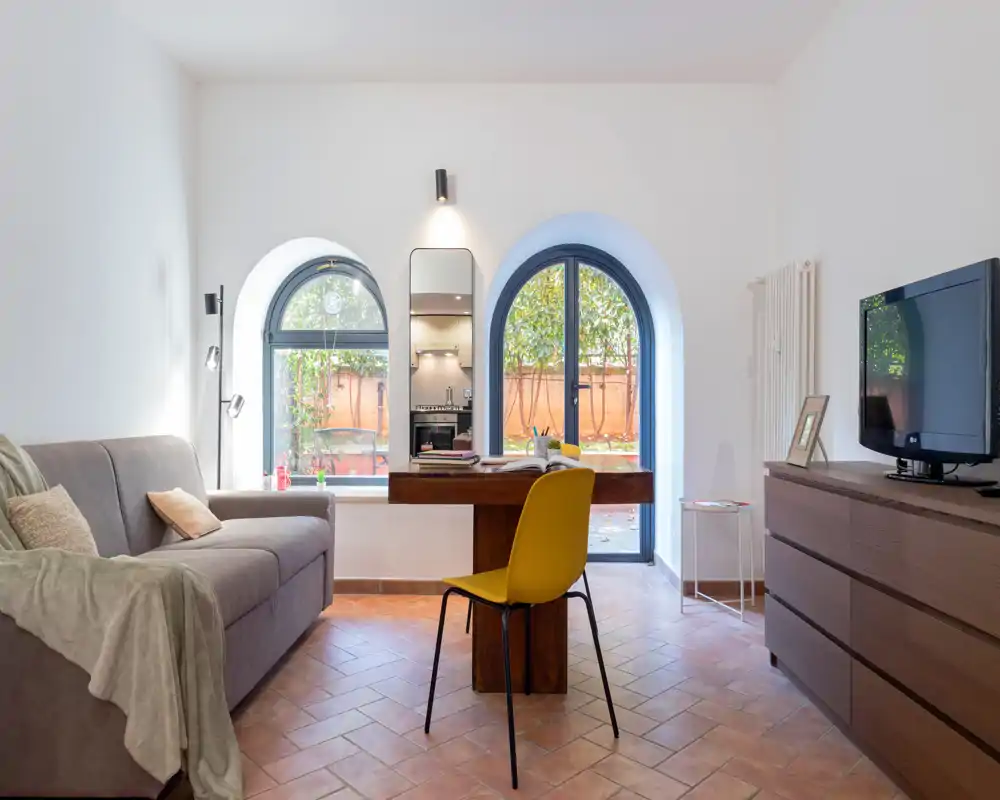 Salon lumineux et élégant avec canapé confortable, bureau en bois, chaise jaune et grandes fenêtres cintrées donnant sur la verdure — parfait pour se détendre ou travailler.