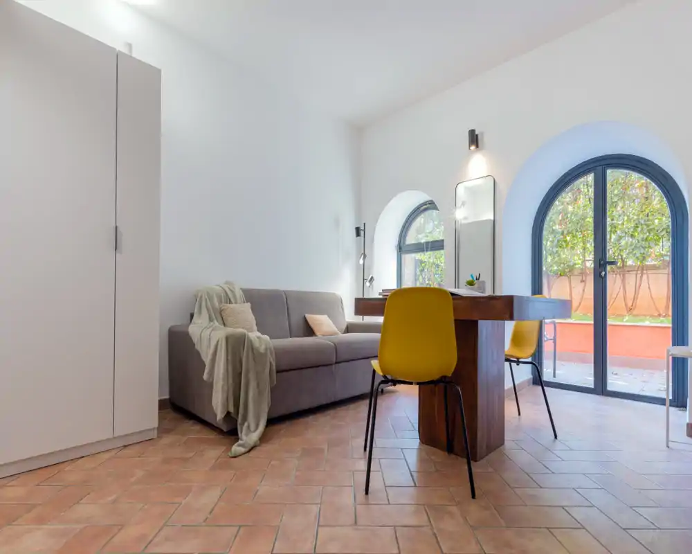 Espace de vie lumineux et moderne avec un canapé gris compact, une table en bois utilisée pour manger ou travailler et des fenêtres en arc donnant sur une petite terrasse. Décor épuré et sol en terre cuite chaleureux.