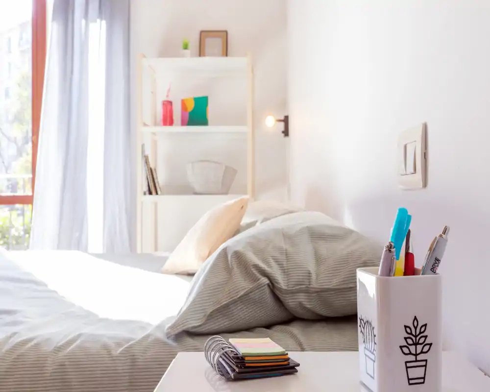 Dormitorio luminoso y ordenado con ropa de cama a rayas, almohadas y una mesita con material de escritura; la luz natural de una gran ventana crea un ambiente fresco y acogedor.