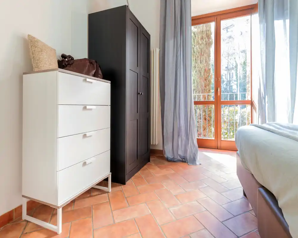 Rincón de dormitorio soleado con suelo de terracota, cómoda blanca, armario oscuro y cama parcialmente visible junto a un amplio ventanal que aporta luz natural.