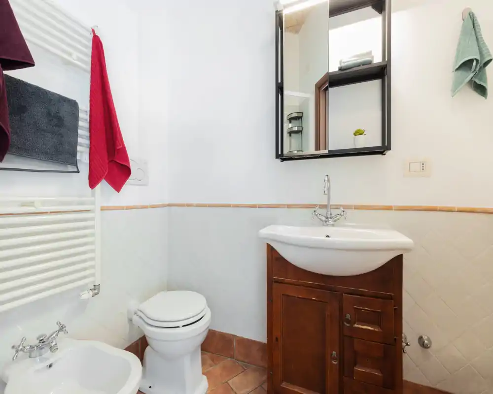 Baño luminoso y bien cuidado con lavabo de cerámica, mueble de madera, bidé y inodoro; azulejos limpios y armario con espejo sobre el lavabo.