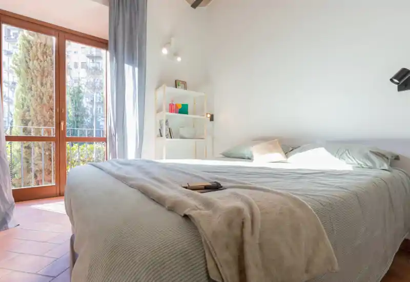 Dormitorio luminoso y aireado con una cama grande vestida con ropa de cama suave, luz natural desde la puerta del balcón y estantería sencilla para almacenamiento.