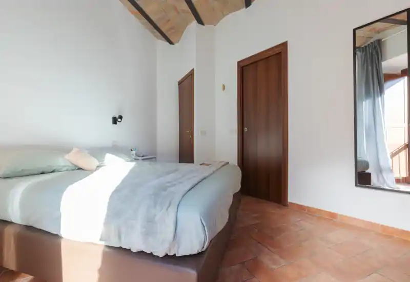 Dormitorio luminoso y aireado con cama doble cómoda, ropa de cama suave y suelo de terracota bajo una bóveda de ladrillo; un gran espejo y la luz natural del balcón realzan la estancia.