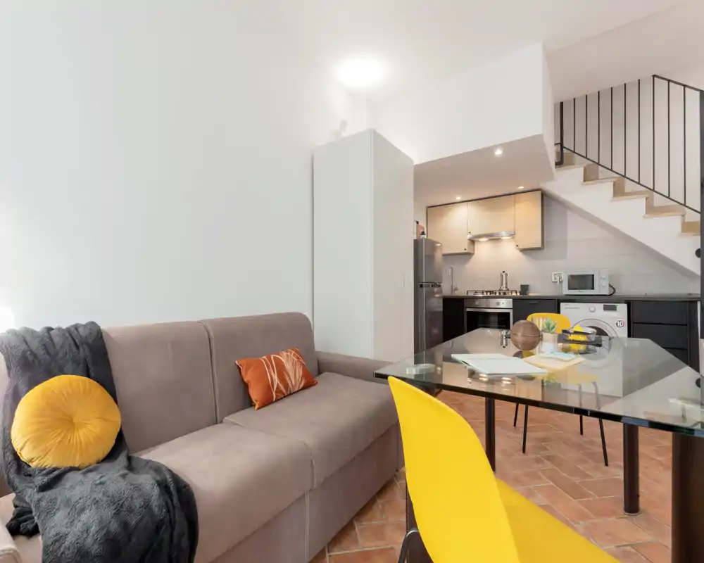 Sala de estar de planta abierta moderna con un cómodo sofá gris, cojines decorativos y una mesa de comedor de vidrio con sillas amarillas; cocina compacta y escalera visibles, espacio funcional y con estilo.