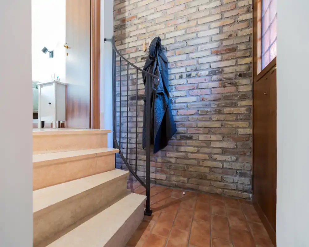 Entrada bien iluminada con suelo de terracota, pared de ladrillo visto y una pequeña escalera que conduce al siguiente nivel; un abrigo cuelga de un gancho junto a la barandilla de hierro.