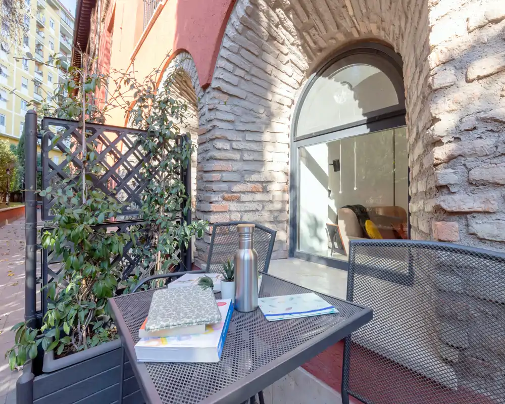 Terraza privada soleada con mesa y sillas de metal, plantas en maceta y un atractivo arco de ladrillo — ideal para café matutino o trabajo al aire libre.