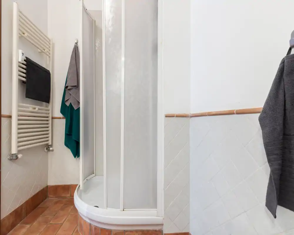 Baño compacto con azulejos y ducha de esquina, radiador toallero de pared y toallas colgadas; espacio limpio y luminoso, apto para anuncios de alquiler.