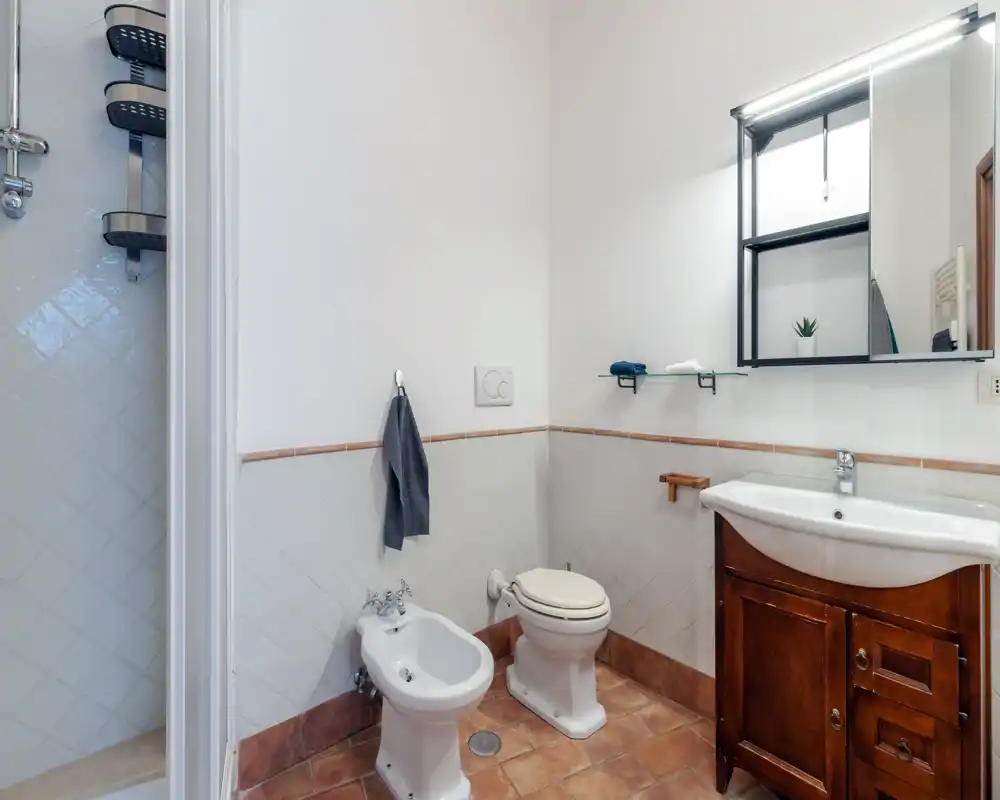Baño limpio y bien iluminado con inodoro, bidé, lavabo en mueble de madera y espejo arriba; suelo de baldosas y plato de ducha visible a la izquierda.