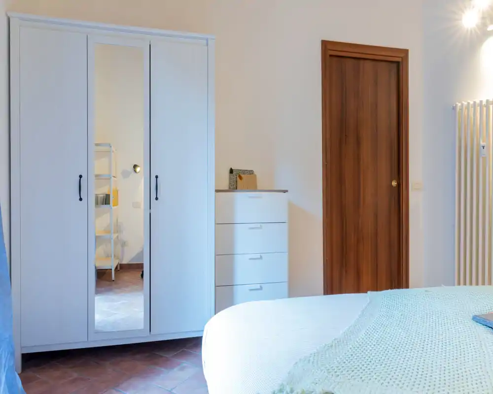 Dormitorio luminoso y ordenado con cama doble en primer plano, amplio armario con espejo y cómoda; colores neutros y agradable luz natural.