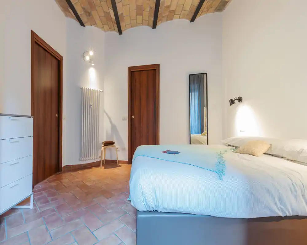 Dormitorio luminoso y acogedor con cama doble vestida con ropa clara, suelos de terracota y un elegante techo abovedado de ladrillo; mobiliario sencillo y espejo de cuerpo entero.