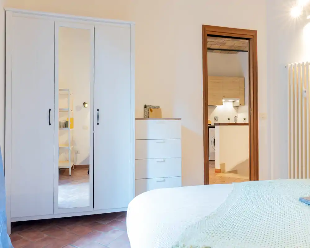 Dormitorio luminoso y de estilo sencillo con un armario claro, cómoda y cama bien hecha en primer plano; tonos neutros y suelo de terracota que aportan calidez.