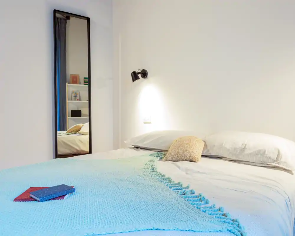 Dormitorio luminoso y minimalista con cama doble bien hecha, manta turquesa suave y lámpara de pared; un espejo alto añade profundidad.