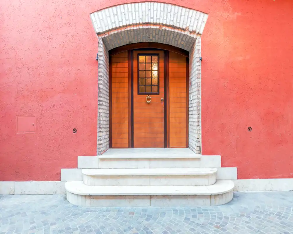 Atractiva entrada exterior con puerta de madera en un arco de ladrillo y tres escalones de mármol desde una calle adoquinada — fachada luminosa y bien compuesta, ideal para destacar el acceso del edificio.
