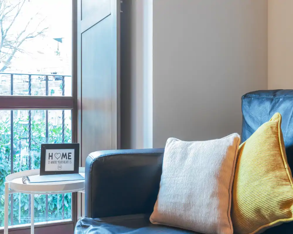 Área de estar luminosa y acogedora con sofá de piel y cojines decorativos junto a una ventana que aporta luz natural.