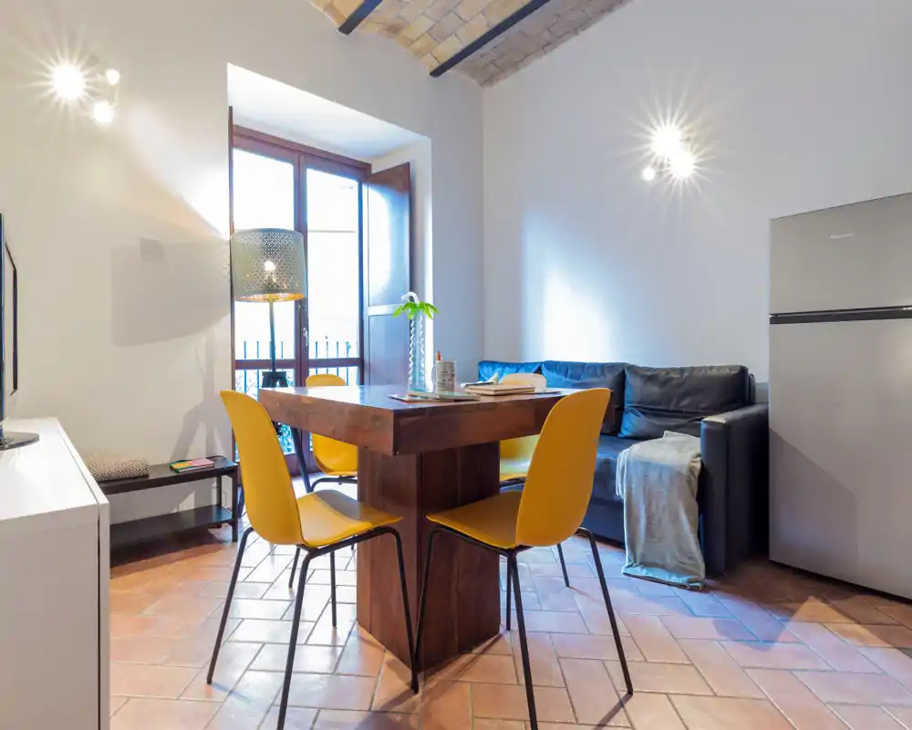 Zona de estar luminosa y con estilo con mesa de madera, cuatro sillas amarillas y un sofá oscuro compacto. La luz natural y las lámparas crean un ambiente acogedor y moderno, perfecto para estancias cortas.