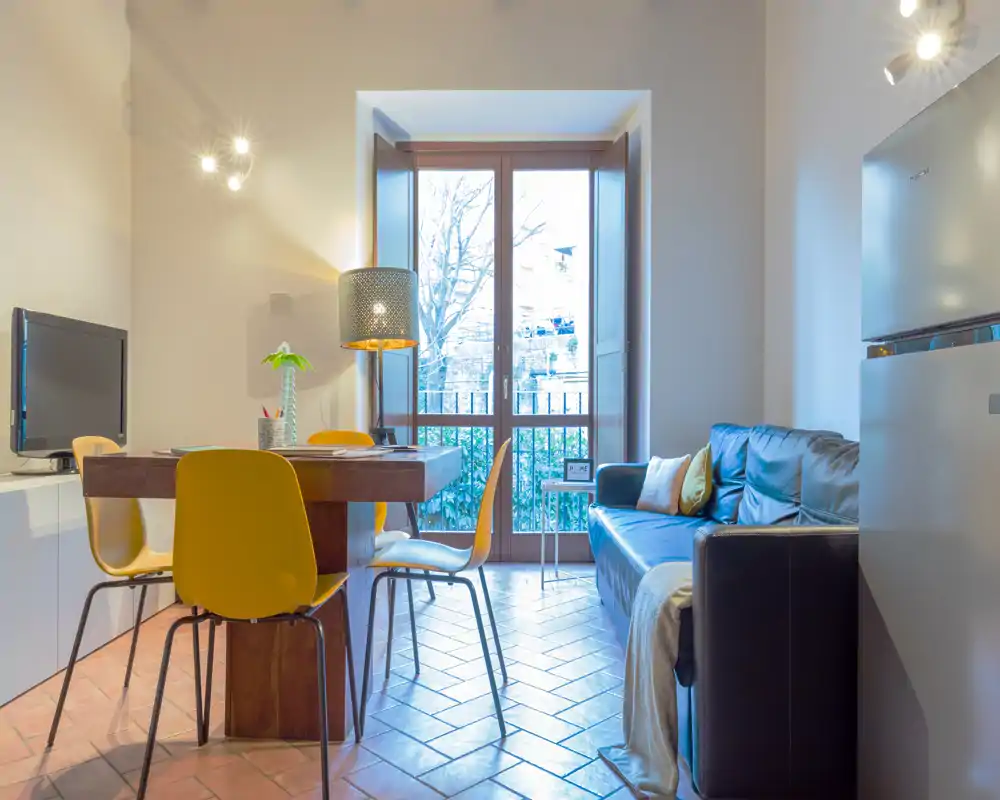 Zona de estar luminosa y moderna con mesa de comedor y sofá junto a una gran ventana, iluminación cálida y mobiliario contemporáneo.