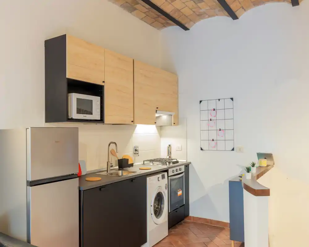 Cocina compacta y moderna con muebles de madera, frigorífico inoxidable, microondas integrado, lavadora y placa de cuatro fuegos — bien iluminada y ordenada para un uso eficiente.