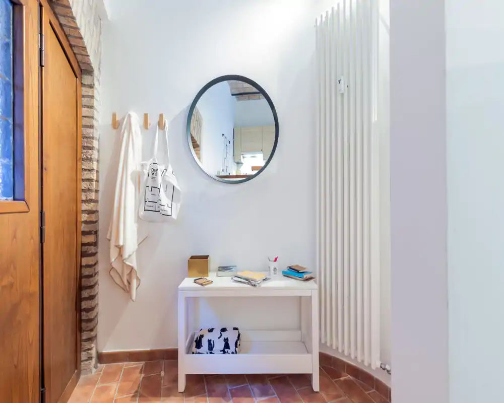 Entrada luminosa y ordenada con una mesa consola pequeña, espejo redondo, perchas con textiles y suelo de baldosas cálido — un espacio de recibimiento acogedor.
