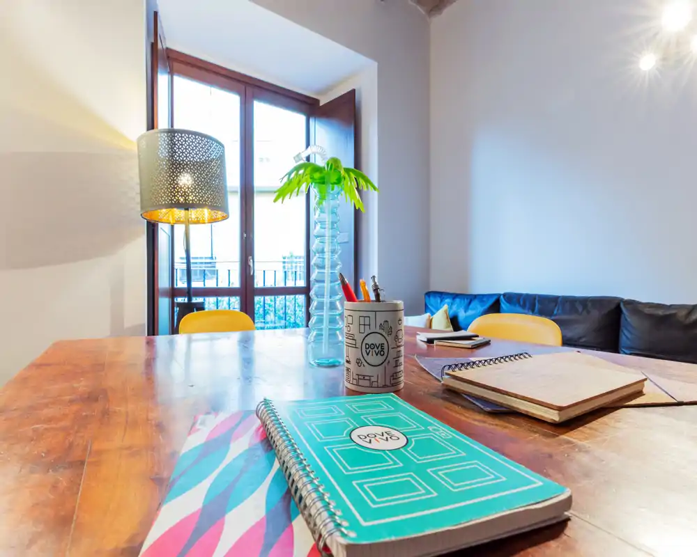 Mesa de madera usada como zona de trabajo informal junto a una ventana luminosa, con cuadernos, un portabolígrafos y elementos decorativos que hacen el comedor acogedor.