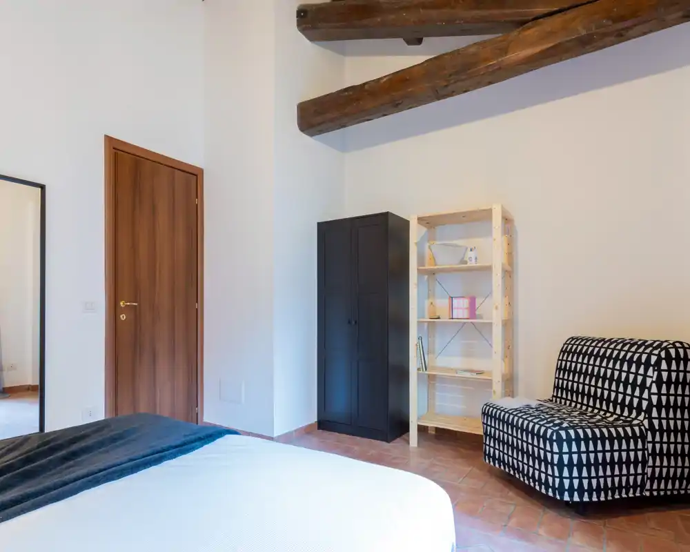 Dormitorio luminoso con cama visible, vigas de madera y suelo de terracota; incluye armario, estantería y butaca estampada.
