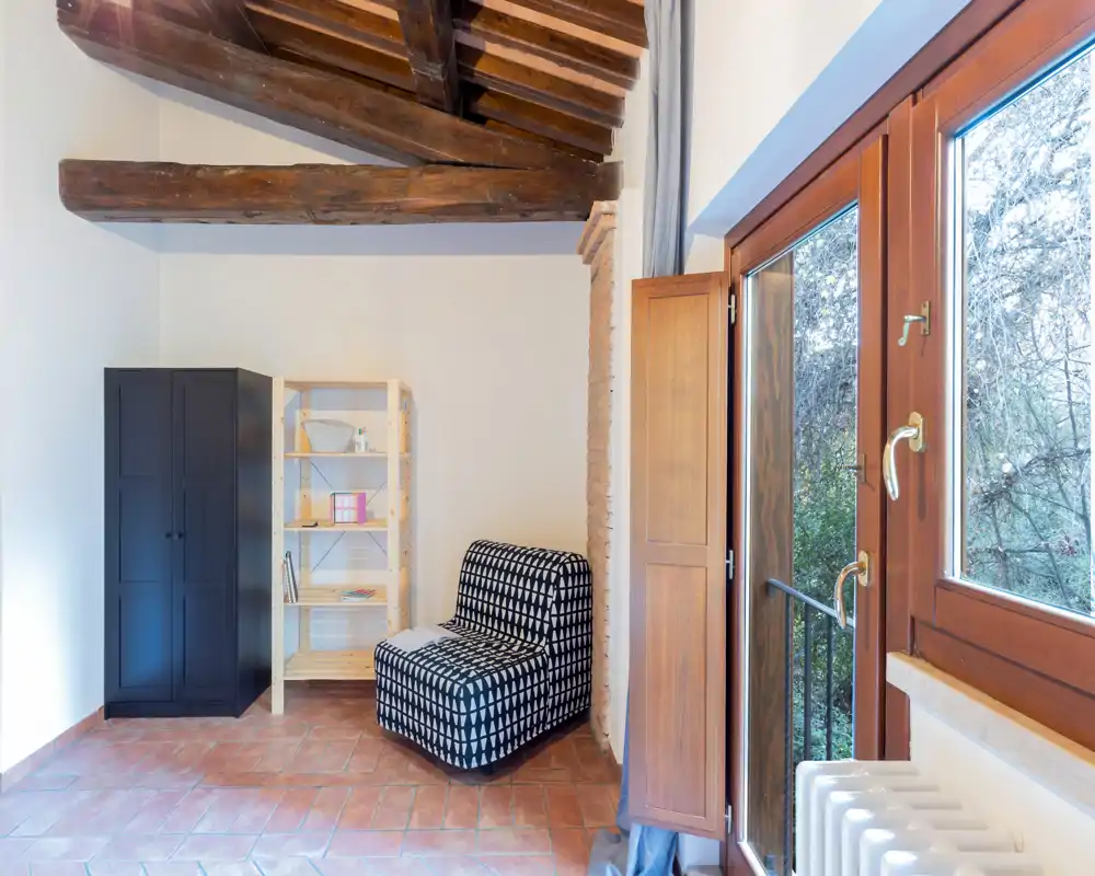 Rincón de salón luminoso con un sillón estampado, estantería abierta y armario bajo vigas de madera; una gran ventana aporta abundante luz natural y una vista agradable.