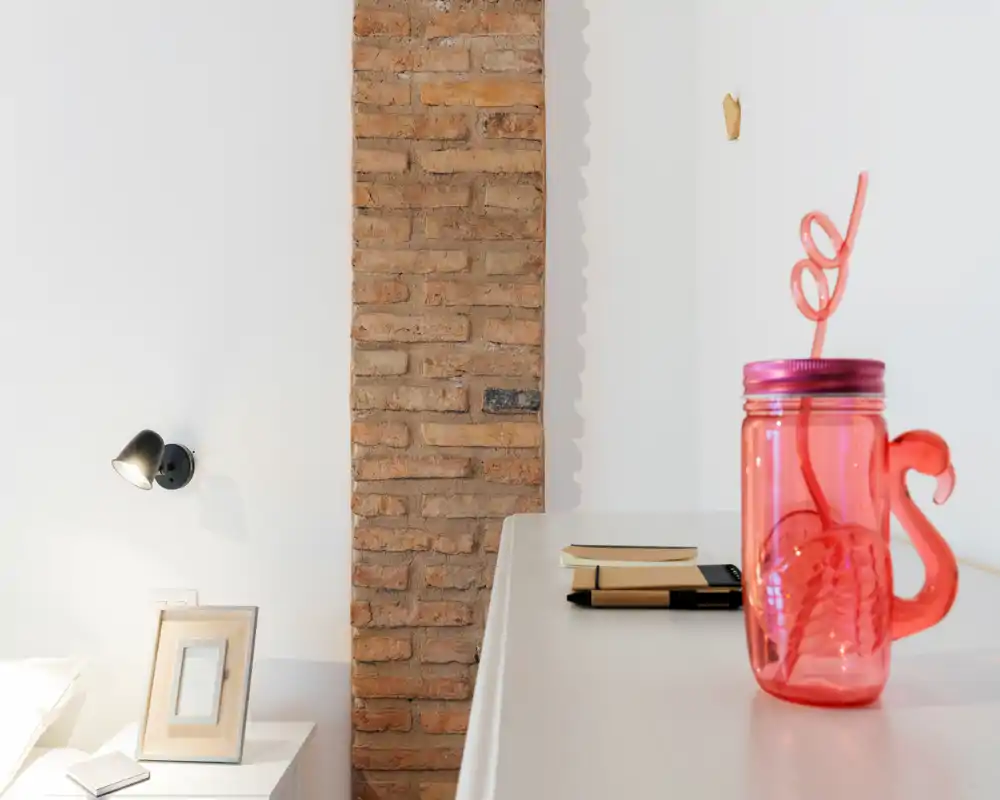 Rincón de dormitorio luminoso y minimalista con ladrillo visto, lámpara de mesita y un tarro rosa decorativo en una superficie cercana. La imagen destaca detalles y luz suave natural.