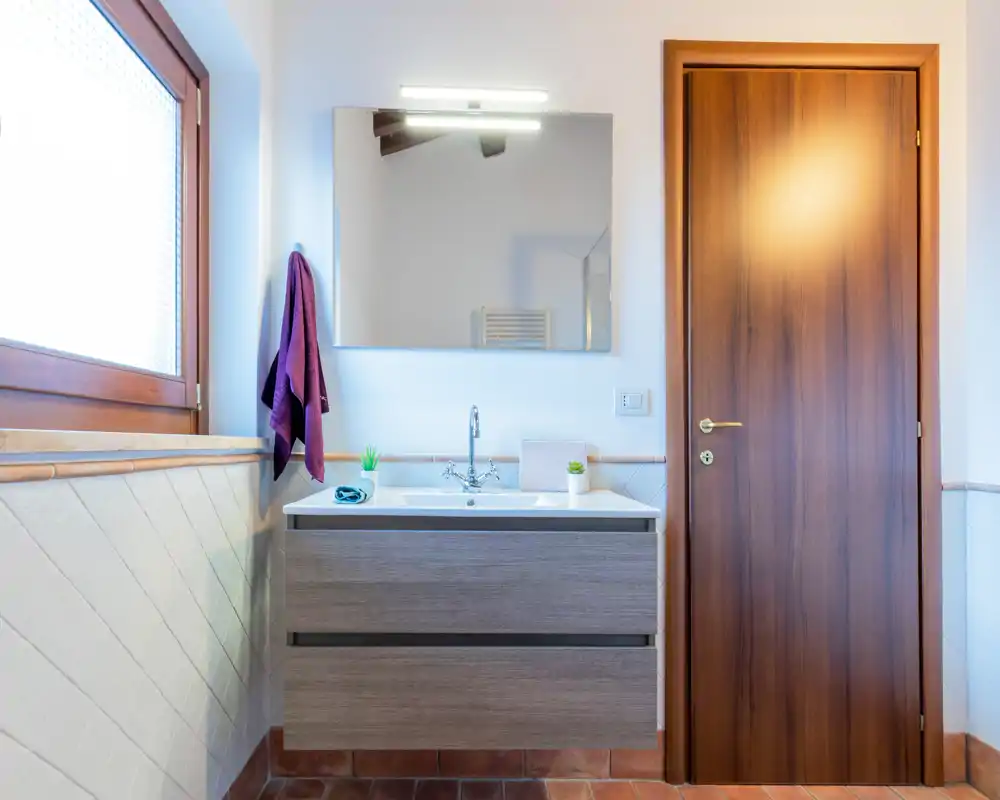 Baño moderno y luminoso con mueble de lavabo suspendido, lavabo rectangular, espejo y puerta de madera. La luz natural de la ventana translúcida y la decoración sencilla crean una sensación de limpieza.