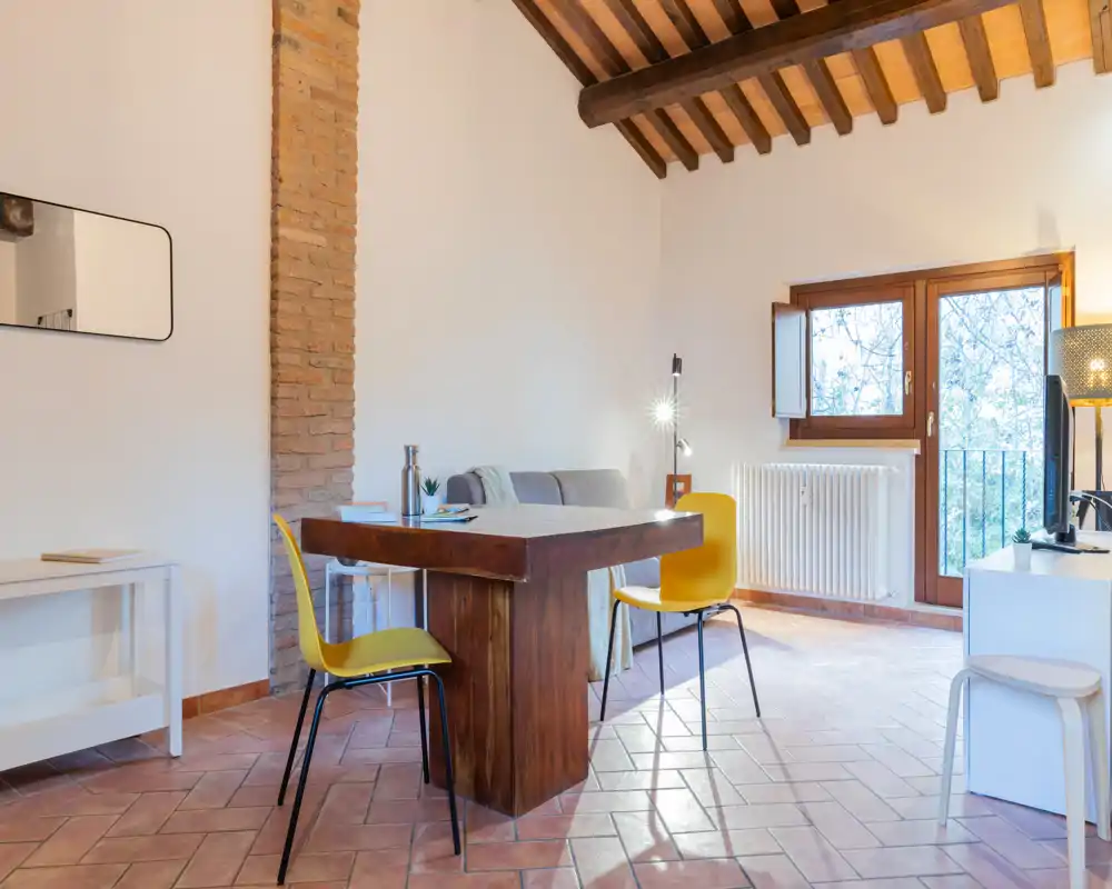 Zona de estar luminosa y aireada con vigas de madera rústicas, mesa central de madera y un pequeño sofá — perfecta para una estancia acogedora con luz natural.