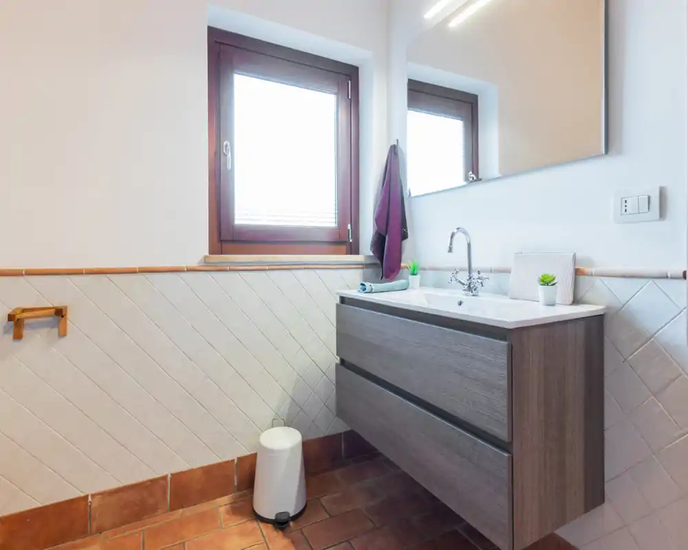 Baño moderno y luminoso con mueble de lavabo suspendido, espejo y ventana que aporta luz natural. Azulejos limpios y suelo de barro cocido ofrecen un aspecto cálido y cuidado, ideal para anuncios.